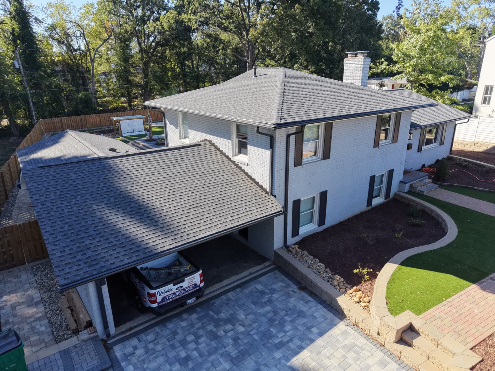 Aerial drone view of a white painted brick two story home with a new charcoal architectural shingle roof, black seamless gutters and downspouts, and the Reliable Roof and Gutter truck parked in the carport