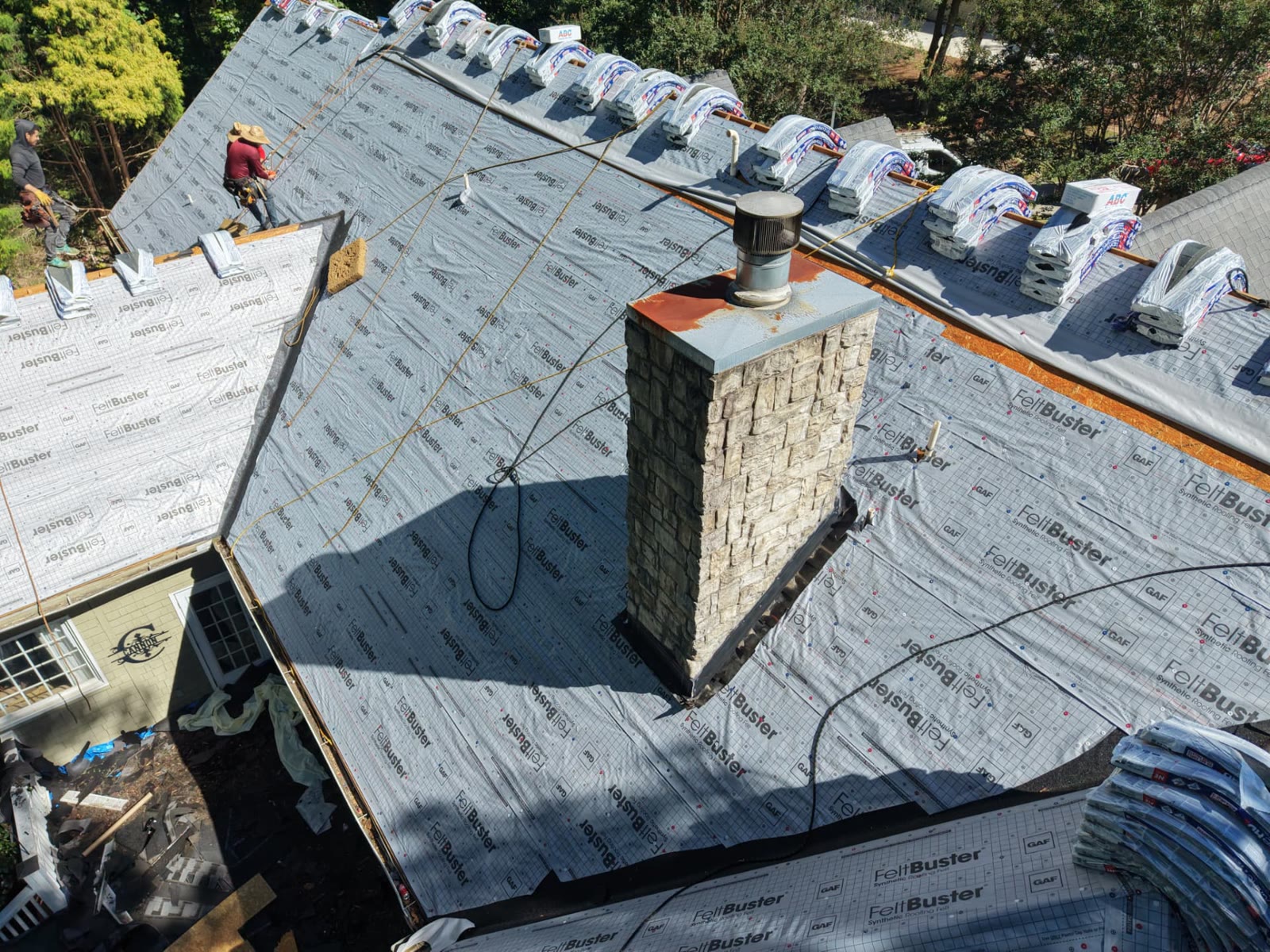 Aerial view of a roof mid replacement with GAF FeltBuster synthetic underlayment, new shingle bundles at the ridge and crew working around a stone chimney