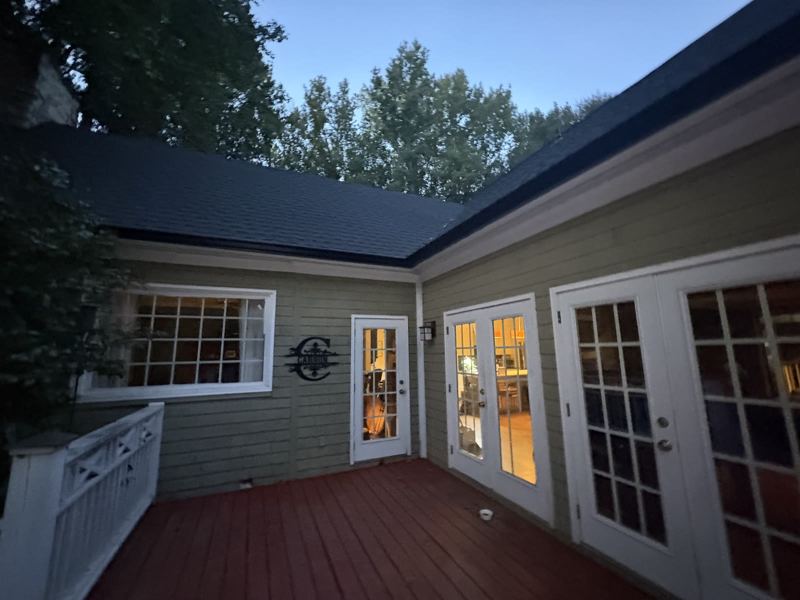 Evening photo of a finished dark charcoal shingle roof on a gray sided home with a red deck and glowing french doors